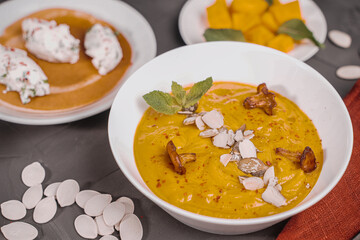Vegetable soup, pumpkin and carrot puree with seeds, mushrooms, chanterelles and mint in a white bowl on a gray background with a burgundy napkin and a black spoon