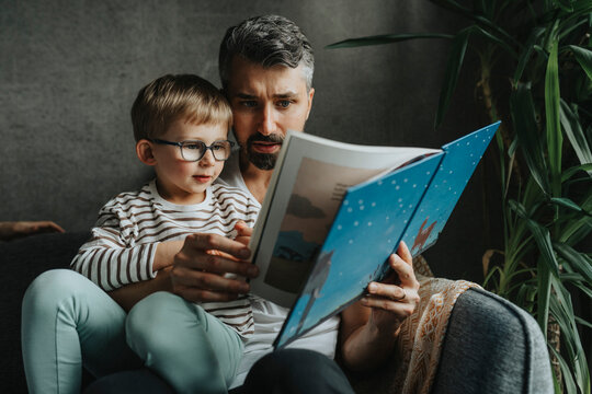 Focused father reading book with son while sitting on sofa at home