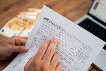 Person reviewing a 1040 tax form with a laptop and euro bills on a wooden desk. Tax filing, personal finance, and accounting concept 