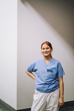 Smiling female trainee standing with hand on hip in front of wall at hospital