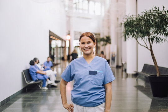 Portrait of smiling female medical trainee standing with hand on hip in hospital lobby