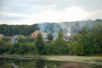 Fototapeta premium Smoke over private houses over the lake and green trees.