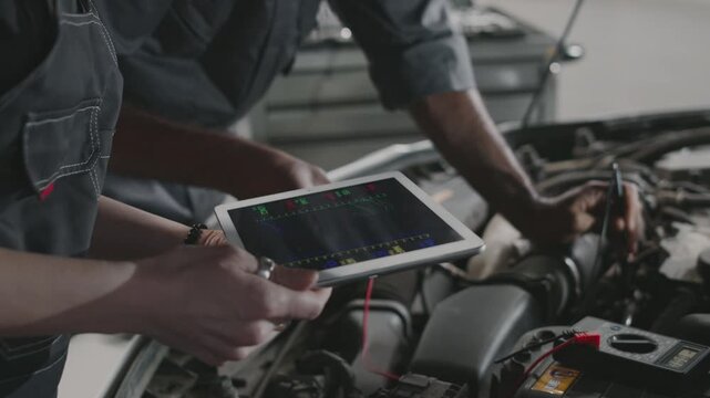 Medium close-up of female technician holding tablet computer wit diagnostic software, and Asian male mechanic checking voltage on starter with amperemeter, while servicing car in garage