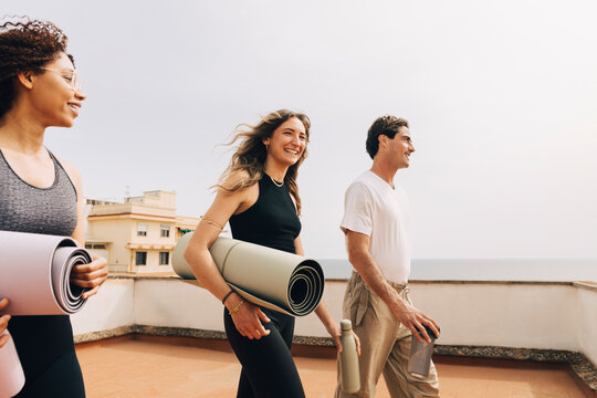 Smiling male and female friends holding yoga mats and walking on rooftop