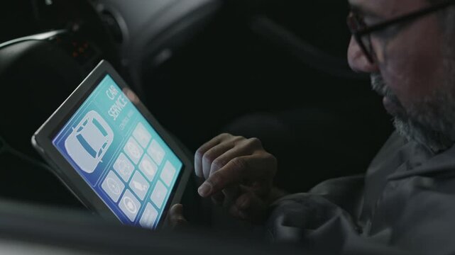 Close-up of middle-aged Asian car mechanic sitting in customer car during complex checkup, testing electric and operating systems, looking at tablet computer with automobile service software