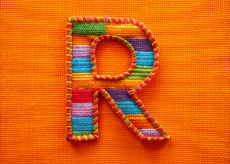 Vibrant orange background highlights a bold, colorful fabric letter R, intricately stitched with multicolored threads, adding a playful touch to any design or message.