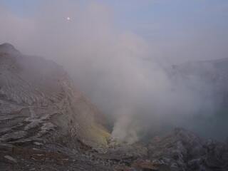 Panoramic view of Mount Ijen, an active volcano in Indonesia, with sulphur smoke rising and fogs laying around. The scene captures the intense atmosphere of this natural wonder.

