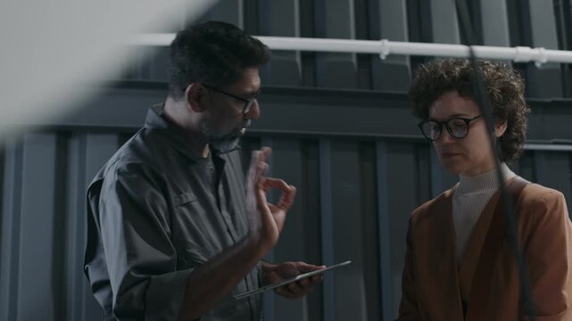 Medium shot of middle-aged Indian technician in grey overalls, with tablet and young Caucasian woman in casual outfit discussing car maintenance work on engine and negotiating repair price