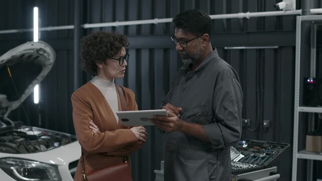 Medium shot of mature Asian technician and Caucasian female customer browsing online for spare parts and supplies for repair on vehicle engine, choosing necessary supplies on tablet, agreeing on price