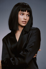 Woman with black hair in a black suit posing confidently with hands on hips against a gray background