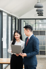 Two business professionals in formal attire discussing work on a laptop in a modern office setting.