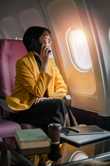 Professional woman in yellow blazer talking on phone and gazing out airplane window, with books and coffee on the table.