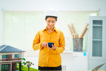 An Asian female architect analyzes blueprints at her desk in the office. She specializes in house design, focusing on roofs and walls, and collaborates with construction contractors in her company.