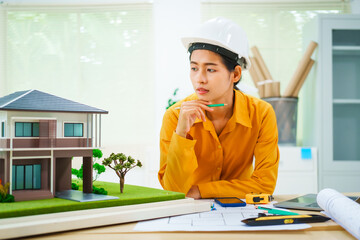 An Asian female architect analyzes blueprints at her desk in the office. She specializes in house design, focusing on roofs and walls, and collaborates with construction contractors in her company.