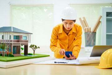 An Asian female architect analyzes blueprints at her desk in the office. She specializes in house design, focusing on roofs and walls, and collaborates with construction contractors in her company.