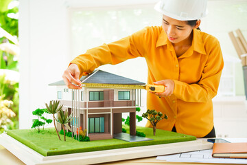 An Asian female architect analyzes blueprints at her desk in the office. She specializes in house design, focusing on roofs and walls, and collaborates with construction contractors in her company.