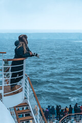 Antarctica - December 26th 2023 - Photo of A23a Iceberg from Cruise Ship Behind Crowd. Passenger With Warm Hat Leans, Holds Camera Looks At Huge Ice © And They Travel