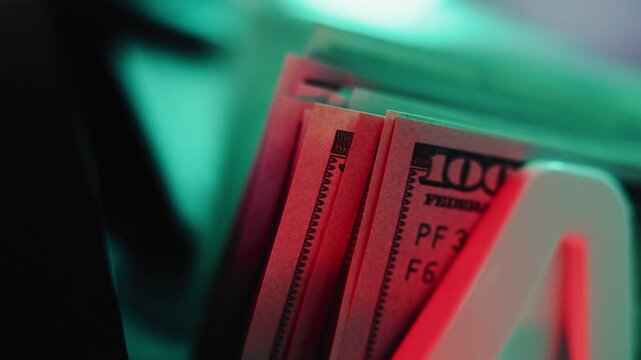 Close up of bill counter machine counting hundred dollar bills for currency exchange. Automatic paper money counting equipment. 100 USD banknotes . Finance and banking. Red and green illumination.