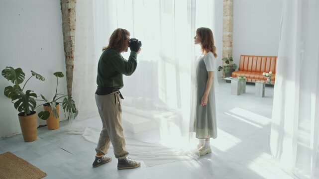 Young female model posing behind sheer curtain in front of professional photographer taking pictures during photo shoot in well-lit modern studio with minimalist interior