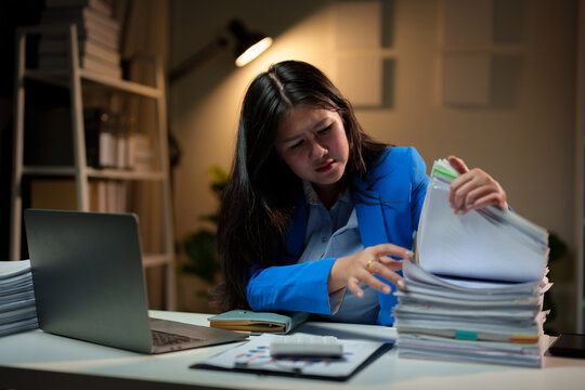 Stressed and tired Asian businesswoman feels a headache while working overtime at night with a pile of documents. Searching, sifting through documents, can't find them. Problems around the office.