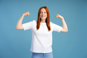 Fototapeta premium Redhead woman showing her strength flexing biceps muscles on blue background