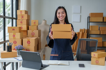 Small business entrepreneur, SME, young Asian woman Freelancer working from home standing holding a box Shipping information on  Laptops online, marketing, packaging, SME, ecommerce concepts.
