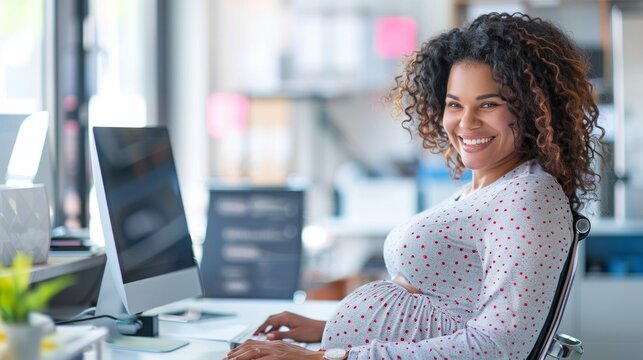 Pregnant biracial businesswoman working at a desktop computer in an office smiling. A woman is sitting at a desk with a computer and a keyboard. She is smiling and she is happy
