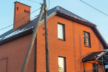 A newly built two-storey red brick house. Brickwork. The roof is made of metal tiles, a chimney for a fireplace on the roof of the house. An old wooden pole of a power line.