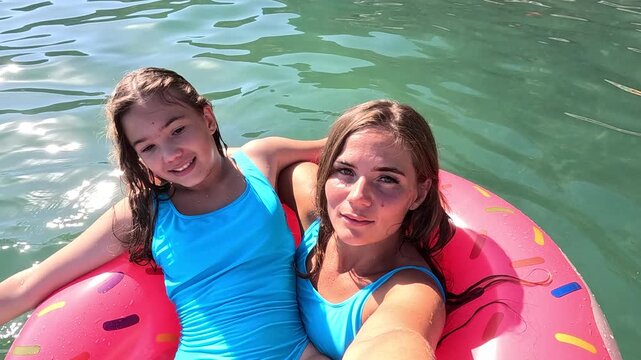 Two young women in blue swimsuits float on a pink donut-shaped inflatable in a body of water. They are smiling at the camera.