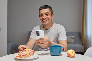 Smiling cheerful positive handsome man sitting at table eating pizza and scrolling his phone in home interior reading interesting post in social network communicating online with friends