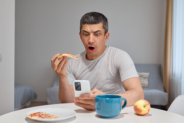 Shocked Caucasian man sitting at table eating pizza and scrolling online using phone in home interior looking at device screen with open mouth being surprised and astonished