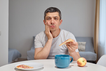 Disappointed upset young man holding piece of pizza feeling teeth pain after eating junk food...