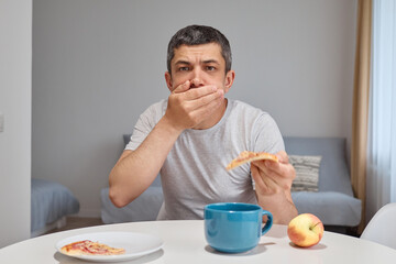 Handsome adult Caucasian man feeling nausea vomit while eating pizza having junk food dinner feels unwell during cheat meal covering his mouth with palm sitting in home interior