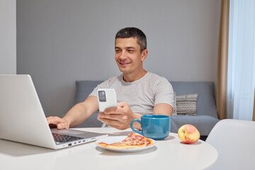Satisfied smiling joyful Caucasian man in casual clothes using smartphone and laptop computer for browsing web pages and internet entertainment sitting at table in home interior