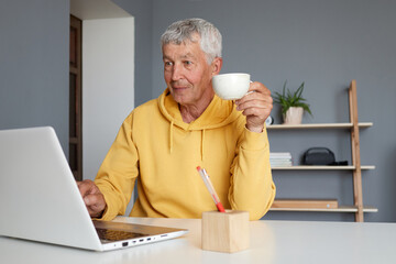 Elderly gray haired man using laptop for work at home and sipping warm drink retired male in yellow sweatshirt relishing coffee while searching information in internet on his computer