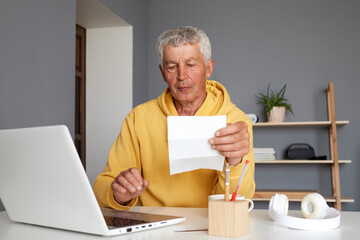 Mature man senior entrepreneur wearing yellow hoodie looking paper bill using laptop computer checking financial invoice or tax document making online payments working at home