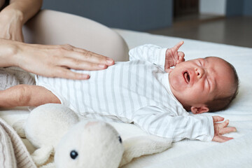 Anonymous unrecognizable mother gently massaging belly of newborn crying baby experiencing colic at...