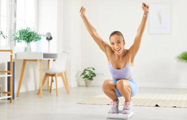 Portrait of happy excited young sporty slim woman measuring her weight standing on scales in the living room at home with hands up rejoicing in successful workouts, dieting and fitness training.