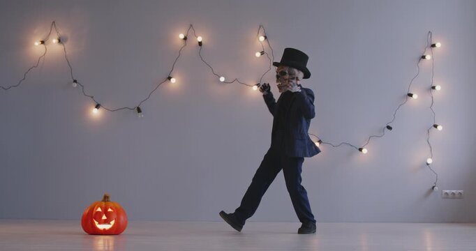 Dancing kid in spooky Halloween costume. Little kid wearing black suit, skull mask, skeleton gloves and top hat dancing in studio with festive lights on gray wall and Jack-o-lantern on floor