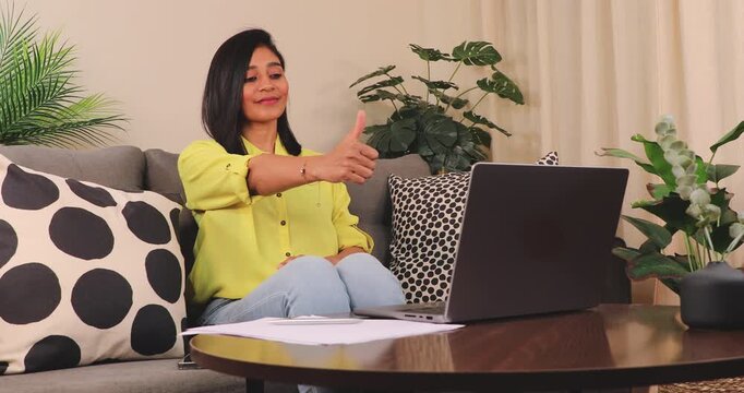 Happy South Asian woman on the sofa looking at a smart laptop and showing thumbs-up