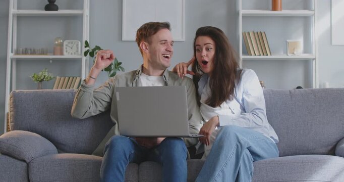 Young couple celebrating online lottery win, reading great news on laptop or getting discount. Family sits on sofa with laptop, is surprised and happy and gives each other high five in triumph.