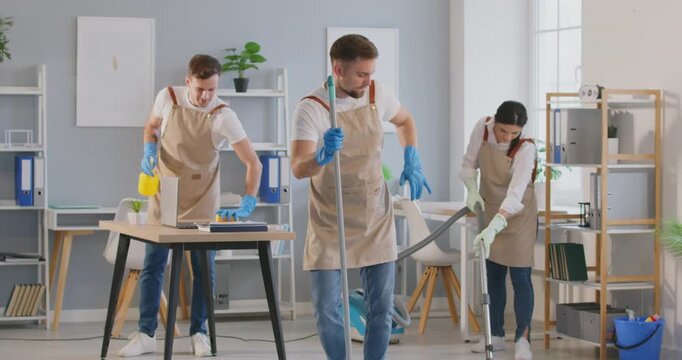 Energetic cleaning service worker dances with mop, adding some fun to routine work. Male cleaner decides to entertain himself and his colleagues while other team members continue to clean office.