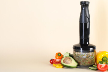 Hand blender with mixture of ingredients and fresh products on beige background, space for text