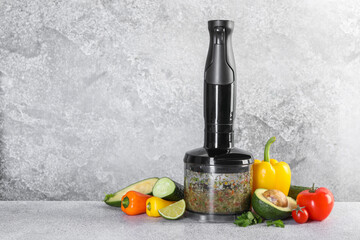 Hand blender with mixture of ingredients and fresh products on light grey table, space for text