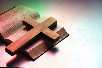 Wooden cross and Bible on textured table in color lights, closeup with space for text. Religion of Christianity