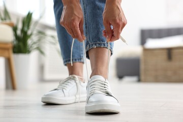 Woman tying shoelace of white sneaker indoors, closeup