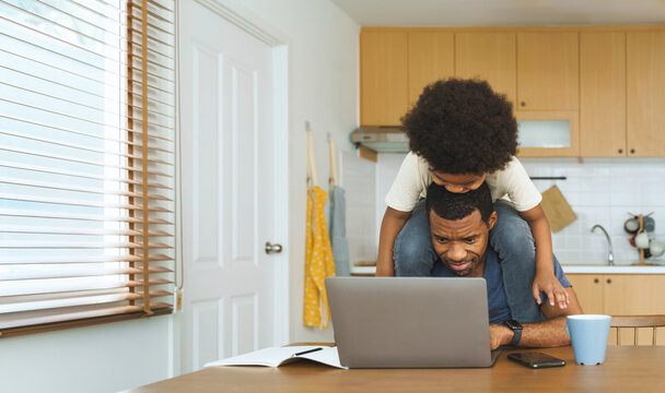 Black African American Father working on laptop with son on his shoulders at kitchen table, playful and determined family bonding.