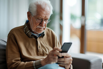 Obraz premium Elderly man sitting on a sofa using a smartphone inside a cozy home. Senior person learning technology and staying connected.