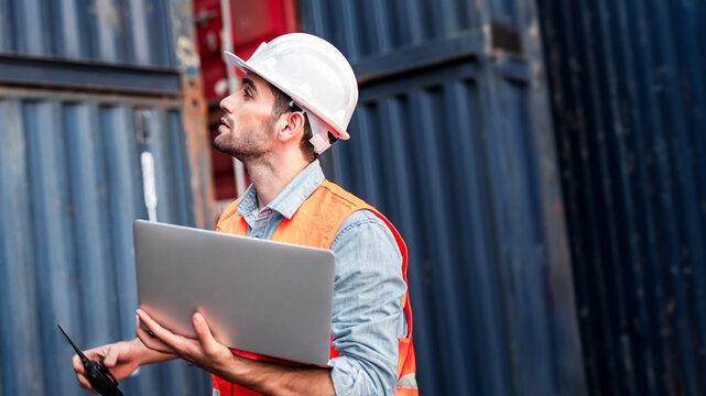 Young confident Caucasian man engineer using laptop computer and wearing white safety helmet and check for control loading containers box from Cargo freight ship for import and export, transport