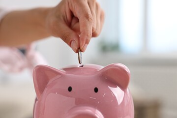 Woman putting coin into pink piggy bank at home, closeup